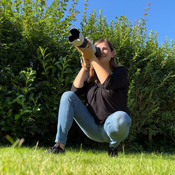 J.M.Bo Die Fotografin kniet im Gras und fotografiert mit einer Kamera in Richtung des Betrachters.